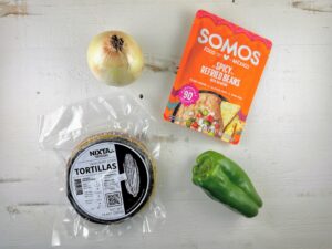 a yellow onion, a green pepper, a package of corn tortillas, and a package of refried beans lay flat against a white wooden background