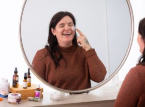 Jaime Sits Before Mirror, Laughing and Applying Moisturizer