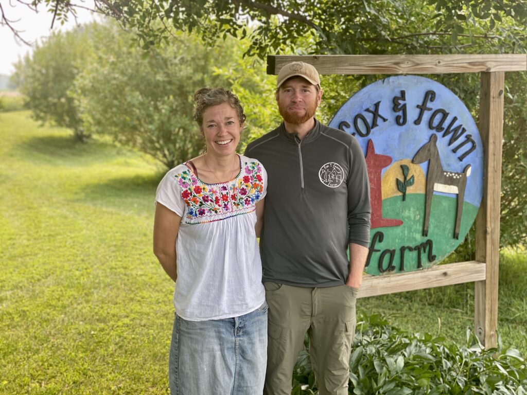 Fox and Farm Farmers Stand in Front of Their Farm Sign